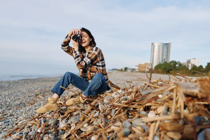 Woman on a rocky beach with a camera, crouching among driftwood in a plaid shirt and jeans, capturing landscape as the calm sea and bright sky frame a casual outdoor photography session.