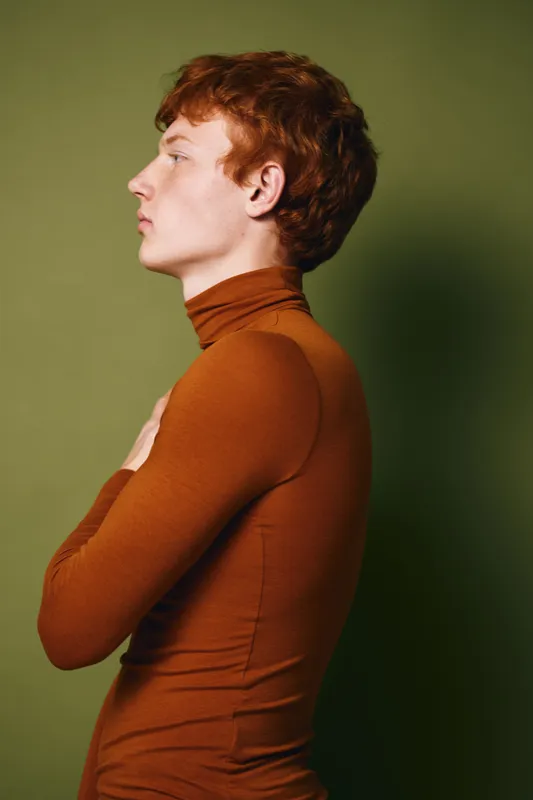 Profile of young man with red hair wearing brown turtleneck standing against green background with folded arms looking forward with thoughtful expression.