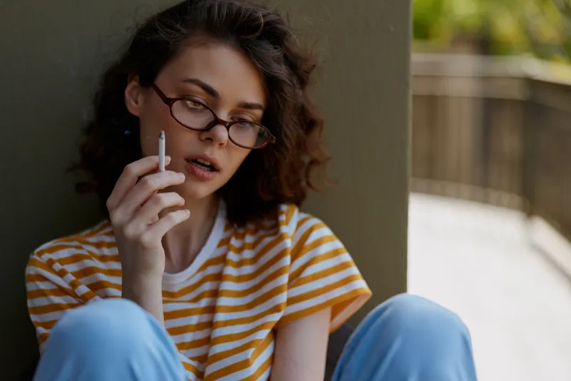 A young woman with curly hair and glasses sits on a balcony, smoking a cigarette while wearing a striped tee and jeans, expressing a contemplative, solitary mood and quiet confidence.