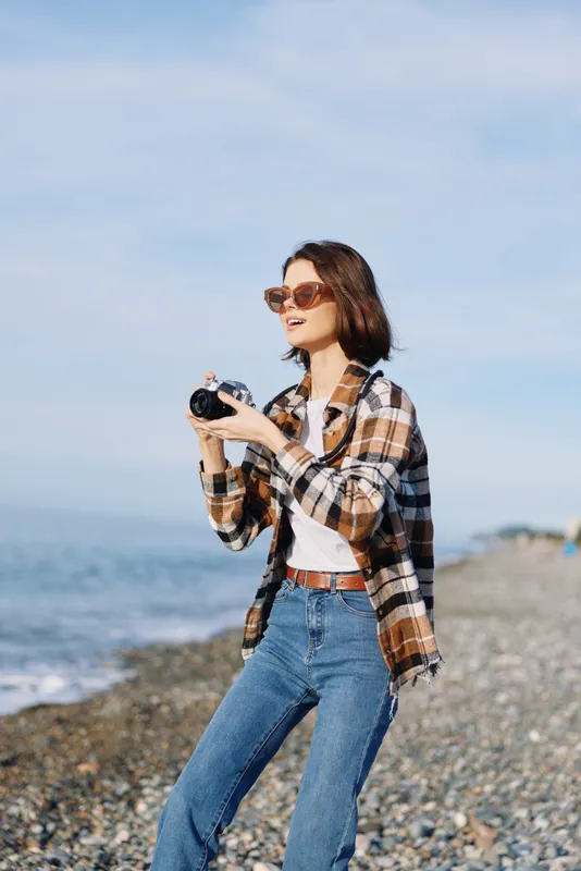 Woman with camera on a rocky beach in casual plaid shirt and jeans, sunglasses, capturing a sunny seascape with a relaxed and stylish photography vibe for travel or fashion shoots.