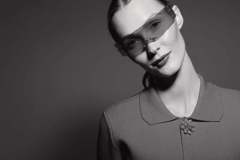 Young Caucasian woman with pale skin wears transparent sunglasses and textured blazer, smiling slightly against dark monochrome background.
