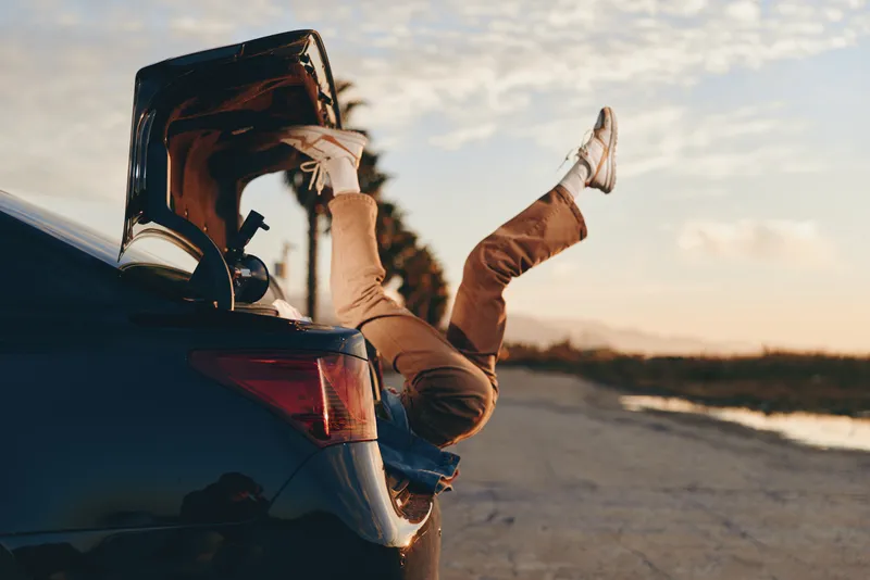 Woman sitting on car trunk at golden sunset, wearing sunglasses and tan jacket, relaxed road lifestyle. Legs out, carefree driving moment on open road. Warm cinematic evening light.