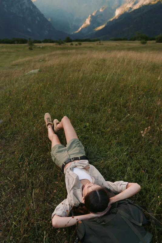 Relaxed young woman lies in a sunlit meadow, feet crossed, looking toward distant mountains with a peaceful smile, a soft breeze rustles wildflowers around her, wearing a light shirt and denim shorts