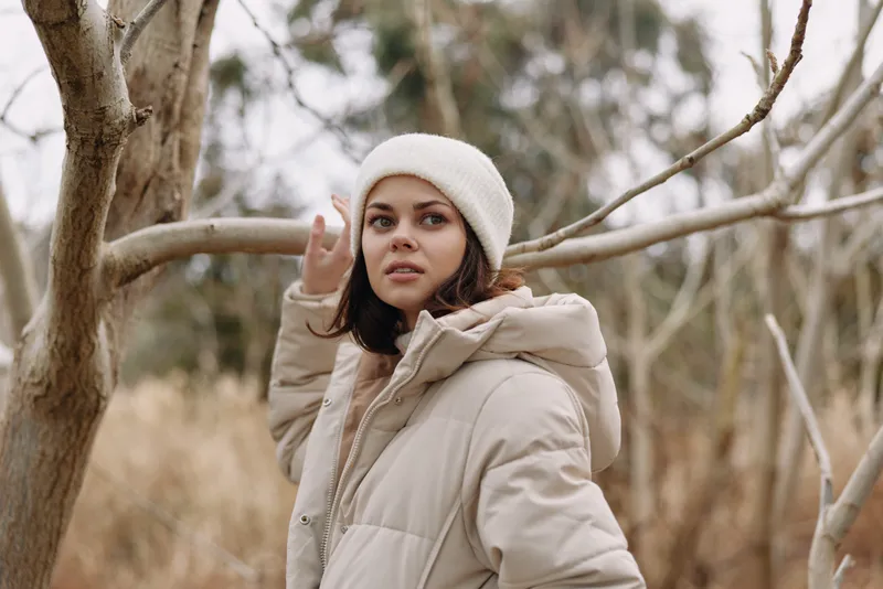 Woman in a beige winter coat and white beanie stands outdoors among bare trees, looking to the side with a calm, thoughtful expression in soft natural light.