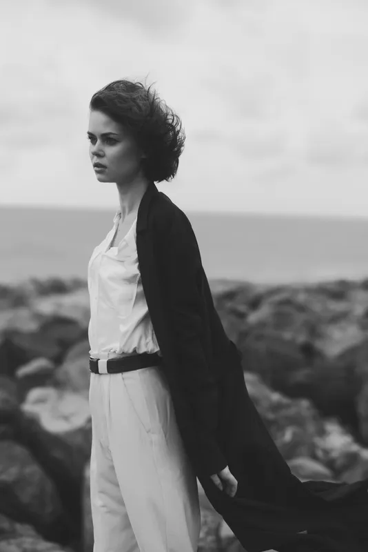 Monochrome fashion scene featuring a woman in a tailored white outfit and flowing black coat on a rocky shoreline, wind-swept hair, serene mood, minimal styling, coastal ambiance, timeless elegance
