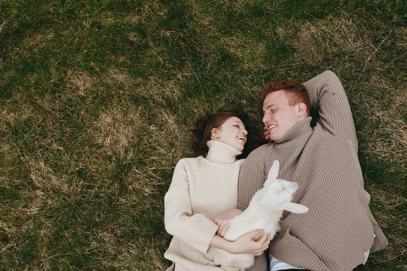 A joyful couple lies on grassy ground with a playful goat kid between them, sharing laughter and warm sweaters. A scene of rural life, companionship, and gentle nature, capturing everyday connection