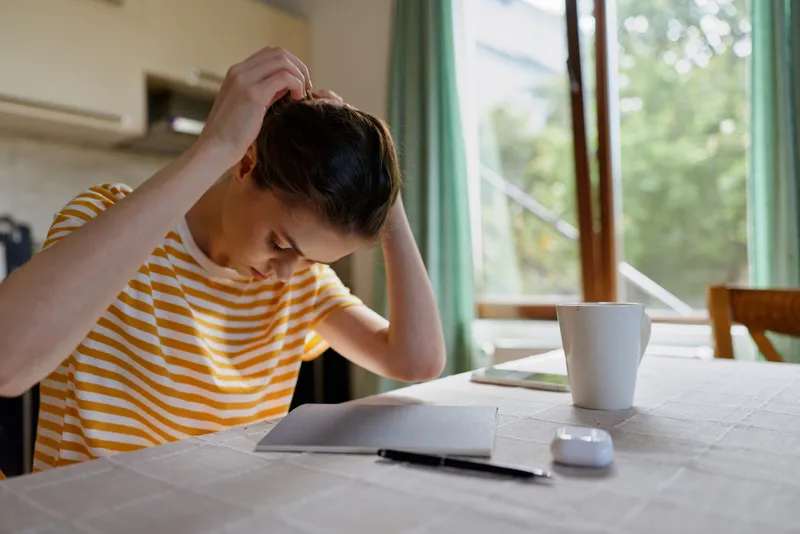 A man in a striped yellow shirt sits at a bright home table, carefully grooming his hair while scattered papers and a mug lie nearby, creating a candid, focused moment that conveys everyday life,