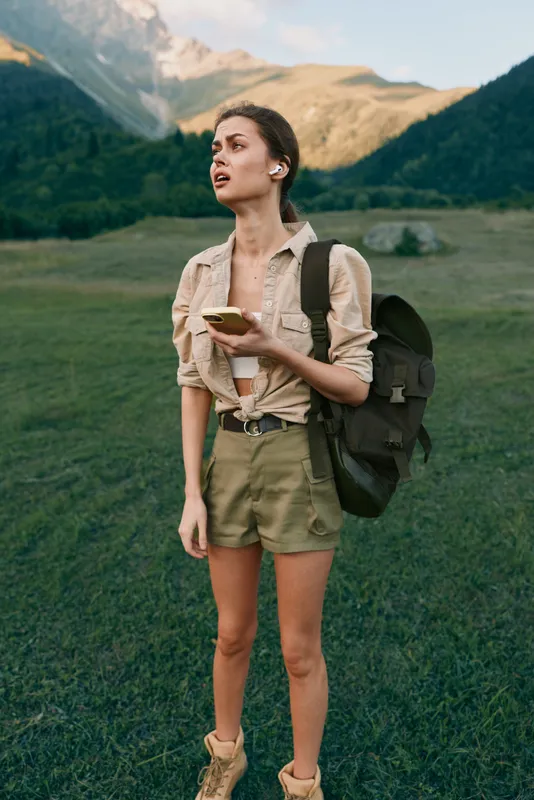 Young woman with backpack standing in a green mountain field holding a smartphone wearing casual summer clothes and wireless earbuds. Outdoor lifestyle and adventure concept.