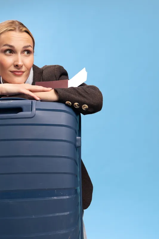 Young woman with suitcase and boarding pass leans on luggage, looking away with thoughtful expression against blue background. Concept for travel, vacation, and journey planning.