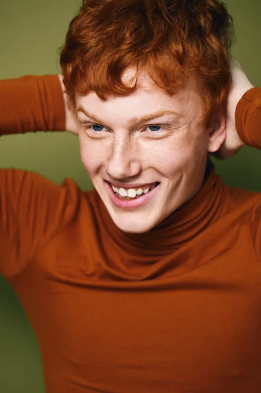 Young man with red hair smiling joyfully wearing brown turtleneck sweater standing against green background. Casual portrait of happy person with hands behind head.