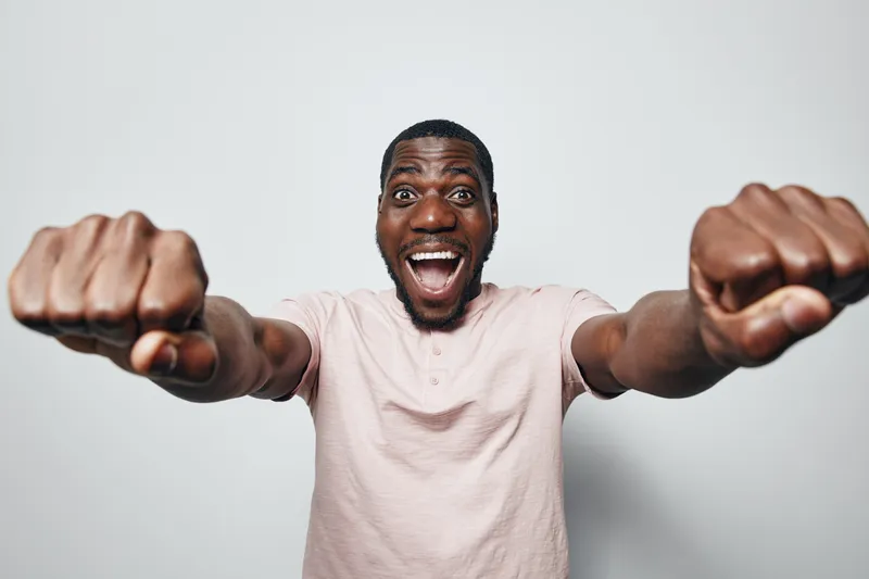 African American man with dark skin wearing light pink shirt smiling excitedly with fists extended forward in studio. Happy male portrait with enthusiasm and energy on white background.