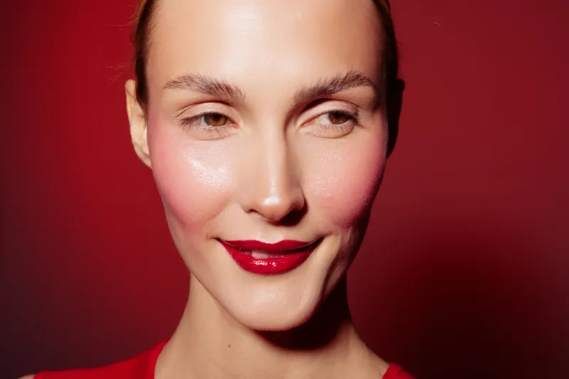 Close-up of a smiling woman with red lipstick and natural skin on red background. Concept of beauty, makeup, skincare, and confident female portrait with studio lighting.