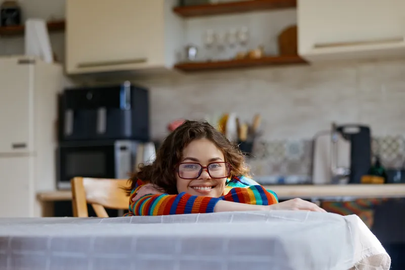 Colorful striped sweater woman rests her chin on a table in a bright indoors kitchen, smiling softly as she leans forward, creating a warm casual domestic moment.