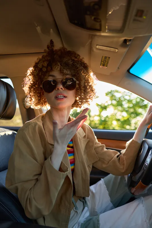 Cheerful woman in a car, wearing sunglasses, lifestyle portrait with retro color, sunlit interior, casual mood, candid moment, warm tones, natural light, relaxed pose, everyday travel