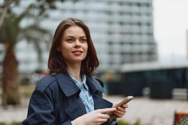 Casual woman outdoors holds smartphone, smiling, standing near an urban backdrop in a denim jacket and shirt, capturing everyday communication moment today.