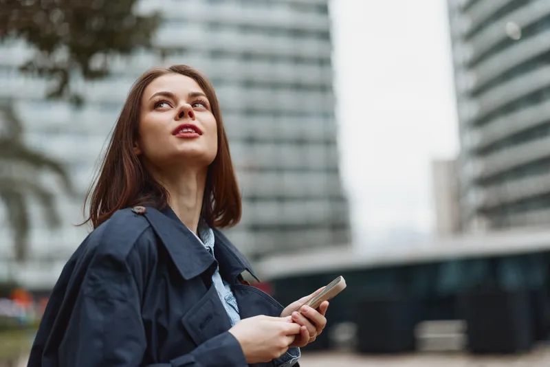 Young woman with long brown hair looks upwards thoughtfully while holding a smartphone in an urban environment during daytime.