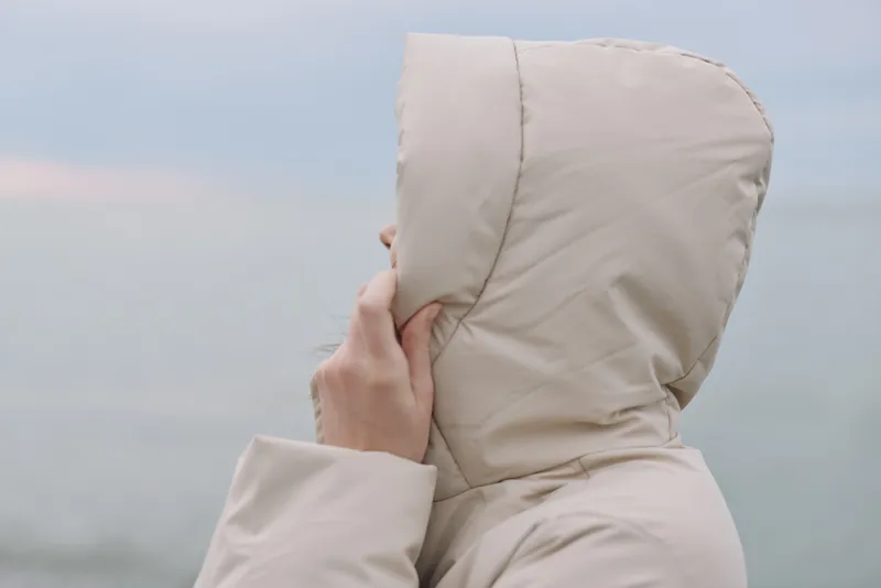 Hooded beige coat on a solitary figure facing the sea, a calm coastal mood with wind and horizon under soft daylight, minimalist outerwear portrait.