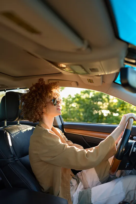 Woman drives a car in daylight wearing a beige jacket with curly hair, retro vibe, focused expression, casual daily commute, warm cinematic lighting, nostalgic mood and bright outdoor scenery.
