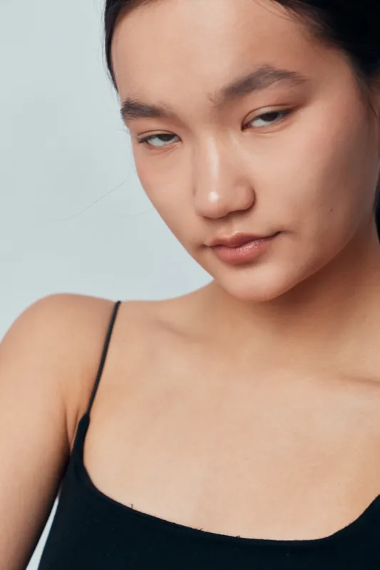 Young woman with long dark hair and natural skin wearing black tank top posing against plain light background with subtle expression and soft lighting in studio portrait closeup.