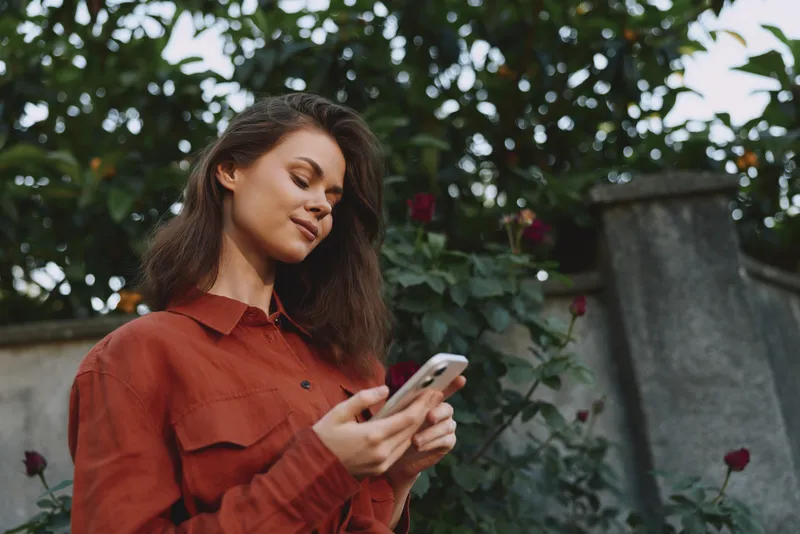 A young woman in a rust shirt checks her phone in a lush garden, capturing a casual moment of everyday life outdoors