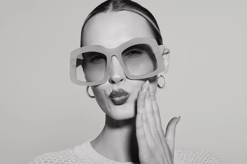 Stylish white woman with pale skin wears oversized sunglasses and earrings, making a playful kiss face. Close-up portrait on plain background. Concept for fashion, beauty, and attitude.