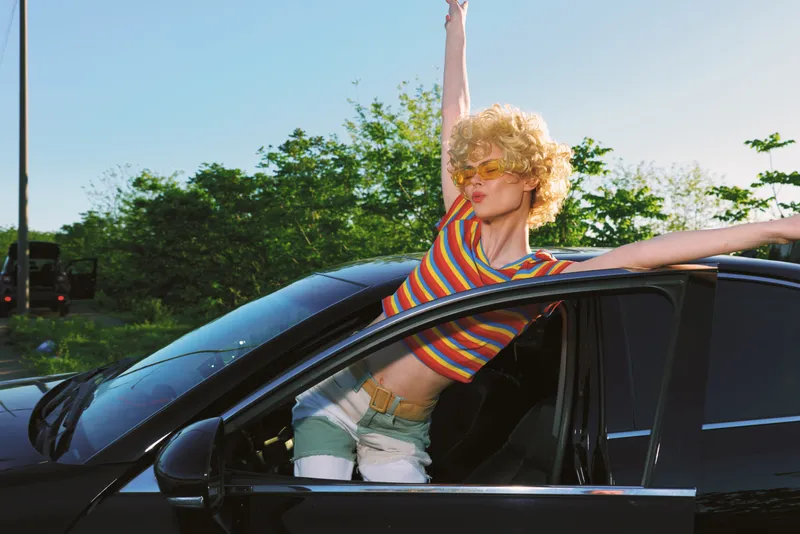 A woman with curly blonde hair leans from a car window in a striped top, capturing a carefree outdoor fashion moment with retro color vibes.