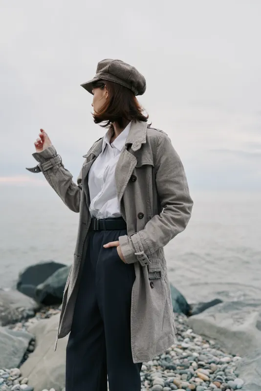 Woman in grey coat and hat stands on rocky shore looking to side, cloudy sky and sea in background. Fashion portrait, autumn style, outdoor mood, casual wear, nature landscape.