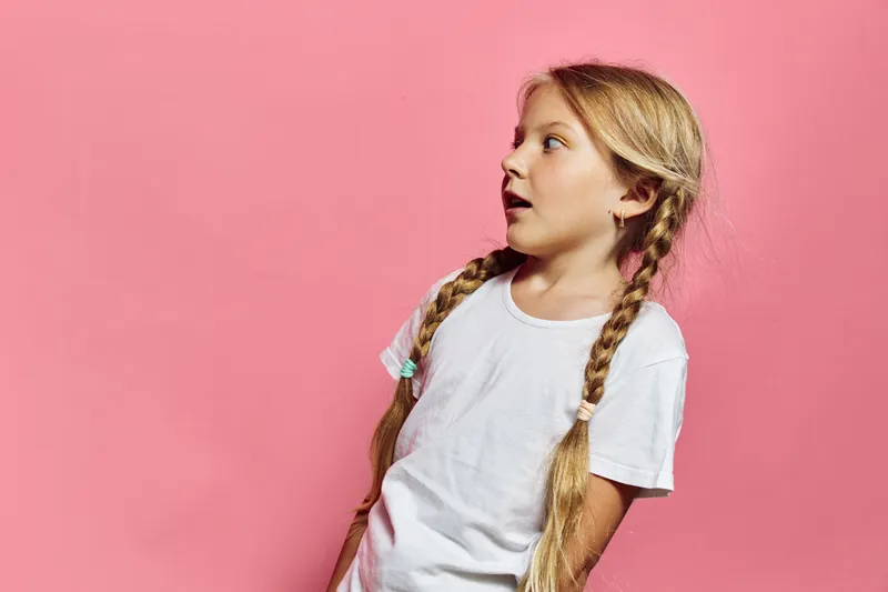 Young girl with long braided hair wearing a white t-shirt looks surprised to the side on a pink background. Studio portrait capturing childhood emotion and curiosity.