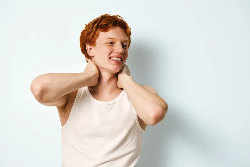 Smiling young man with red hair wearing white sleeveless shirt standing against white background touching neck with both hands in relaxed pose.