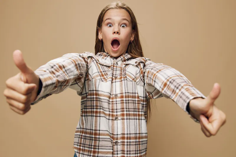 Girl with long hair in plaid shirt shows thumbs up with surprised face expression on beige background for success, approval and positive emotion concept