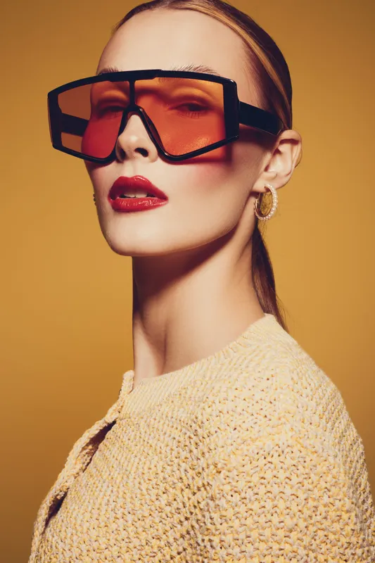Stylish woman with slicked back hair wears oversized orange sunglasses and gold hoop earrings. She poses confidently against a warm yellow background in a textured beige sweater.