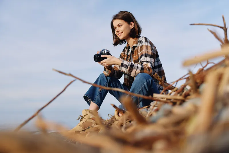 Woman photographer crouches on driftwood by the shore, holding a camera and scanning the scene for the perfect shot as waves roll under a blue sky above the calm coastline.