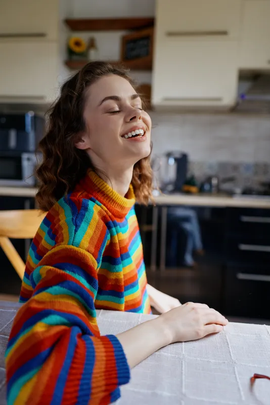 Woman enjoying a joyful moment at home, leaning on a table in a colorful striped sweater, smiling with a bright kitchen backdrop, lifestyle image capturing comfort and relaxation.