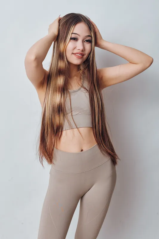 Young woman with long straight hair wearing sport outfit posing with hands on head against white background. Fitness, beauty, health, confident model smiling for portrait photo.
