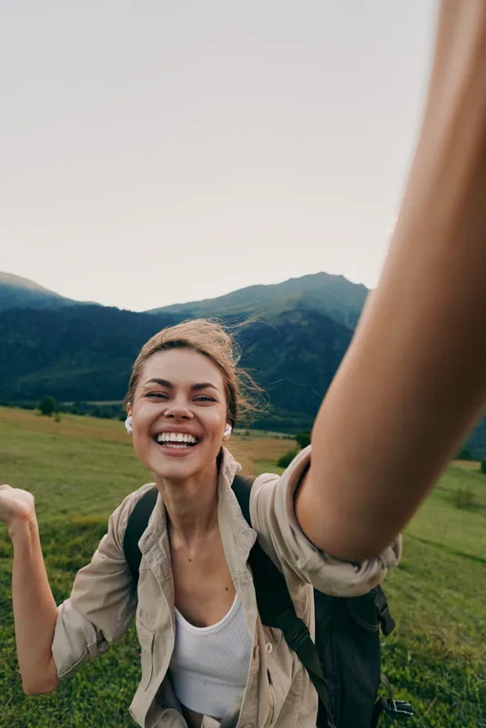 Cheerful woman outdoors, smiling at the camera in a sunlit field, casual jacket, backpack, gentle breeze, natural colors, lush landscape