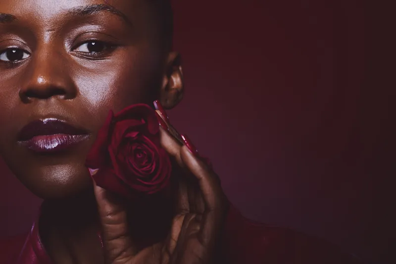 Close-up of a dark skinned African woman with smooth skin holding a red rose near her face. Concept of beauty, elegance, floral, and natural skincare on dark background studio.