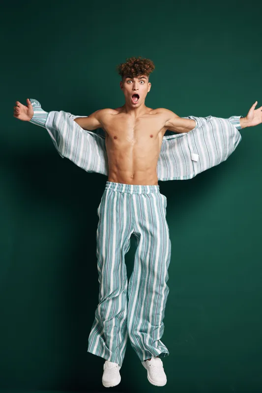 Young man jumping with open arms wearing striped pants and matching open shirt against green background. Shirtless male model with curly hair and surprised expression in studio.