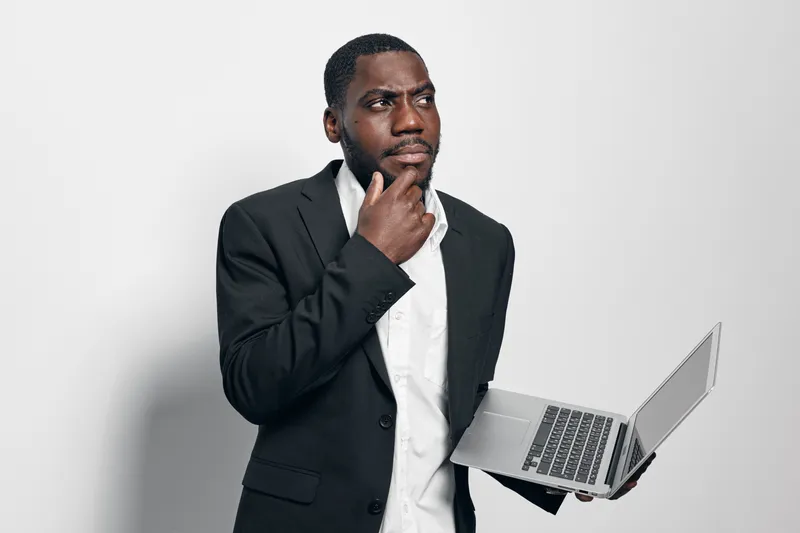 Black man with dark skin in business suit holding laptop and thinking with hand on chin isolated on white background. Professional African American male in formal wear with technology device.
