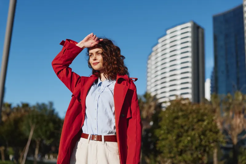 Young woman with curly hair wearing a red coat shields her eyes from the sun in an urban park, projecting confidence and chic street style.