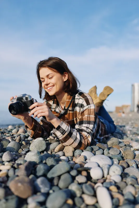 Woman photographer lying on a pebble beach, smiling while capturing images with a camera, wearing a plaid shirt, blue sky and joyful outdoor photography moment