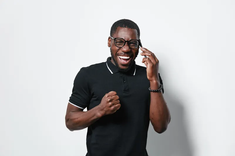African American man with dark skin and glasses smiling and talking on smartphone, wearing black polo shirt, expressing happiness and excitement in studio with white background.