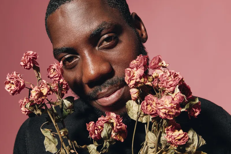 African man with dark skin holding dried pink flowers in creative conceptual style on pink background. Portrait with artistic expression and floral elements in studio lighting.