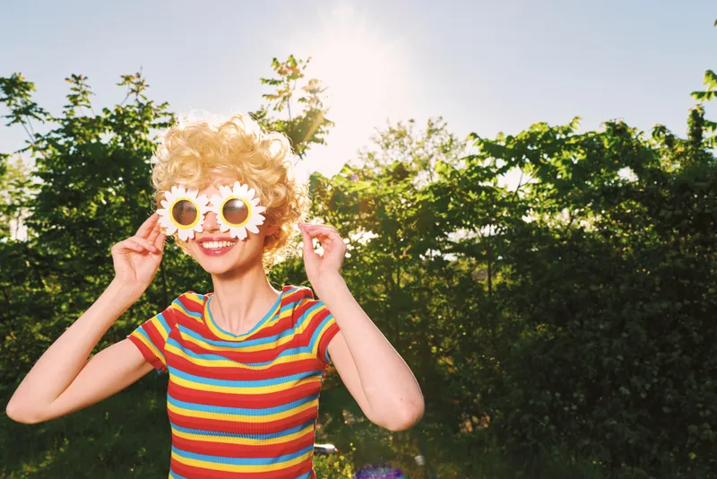 A playful woman in a striped colorful shirt wears quirky flower sunglasses in a sunlit park, capturing a retro lifestyle moment of joy, spontaneity, and outdoor fun, and bright color.
