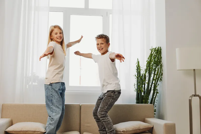 Happy children jumping on sofa in bright living room, smiling and playing together. Concept of family fun, childhood joy, and indoor activity.