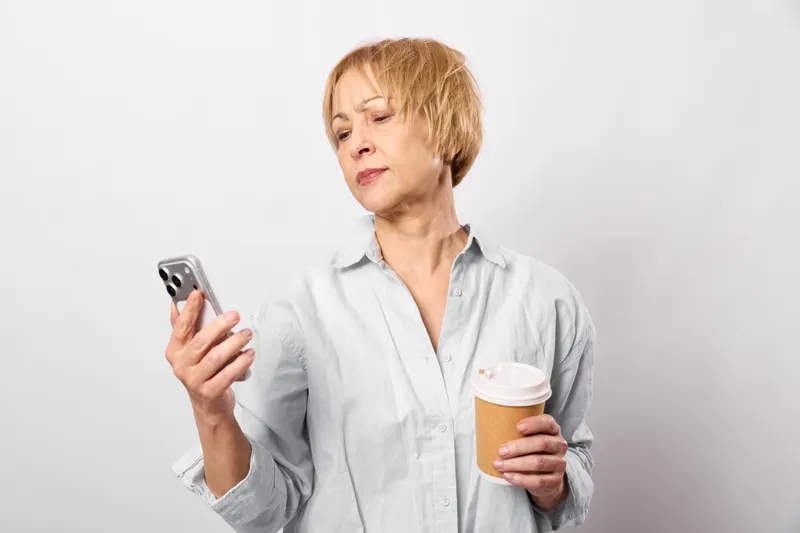 Elderly woman holding smartphone and coffee cup indoors, studio portrait on white background, thoughtful expression, relaxed posture, natural light, copy space
