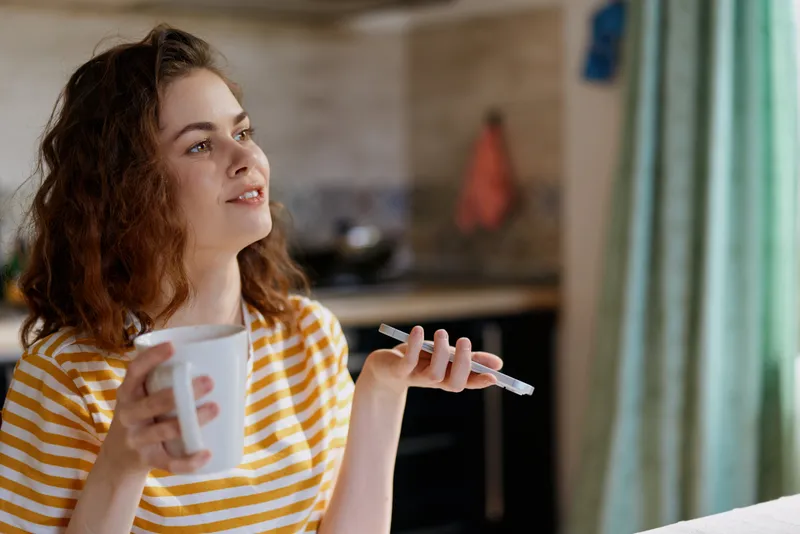 Young woman with curly hair sits in a sunny kitchen wearing a striped shirt, holding a mug in one hand and a pen in the other, smiling softly as she plans ideas for the day