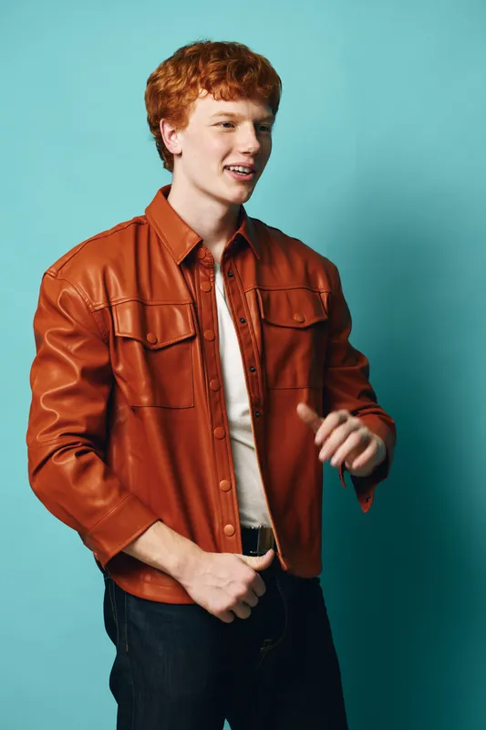 Young man with red hair wearing brown leather jacket and white shirt standing against blue background looking sideways and smiling with casual posture and style.