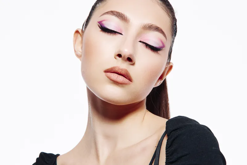 Young woman with creative pink and purple makeup looks downward with closed eyes. Studio portrait on white background. Concept of beauty, fashion, and creative cosmetics.