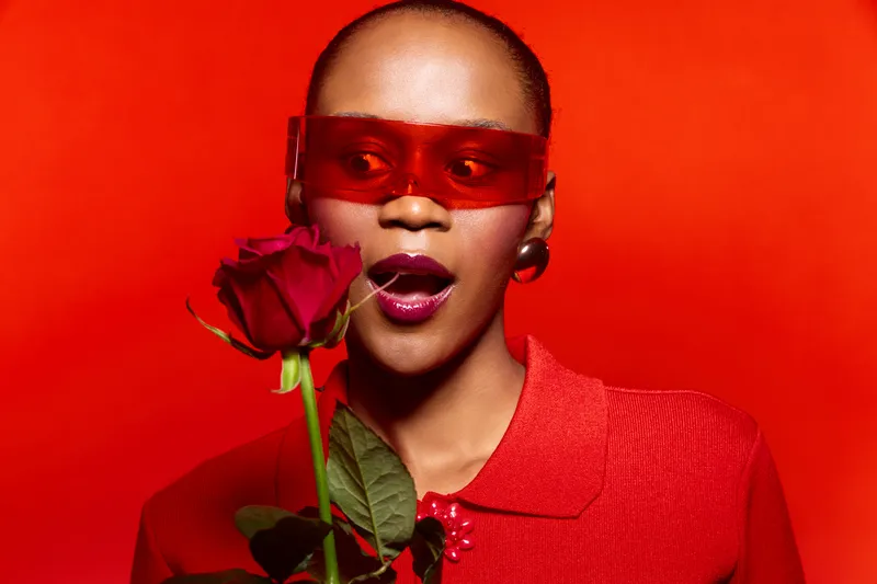 Portrait of a dark skinned African woman with short hair wearing red sunglasses and a red shirt holding a red rose. Studio shot with vibrant red background and fashion concept.