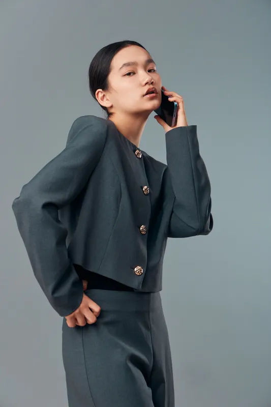 Young woman in stylish suit talking on phone standing indoors with serious expression. Elegant business fashion with minimal makeup and dark hair against gray background.